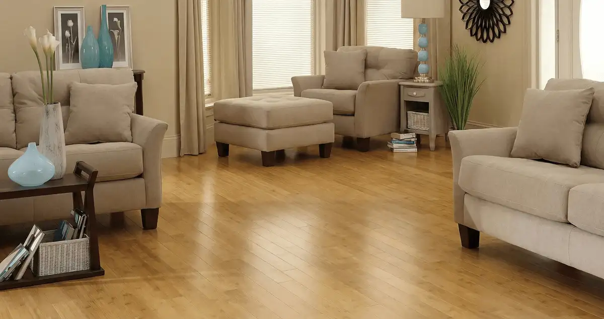 A living room with professionally installed bamboo flooring, beige sofas, a matching ottoman, a side table with a blue lamp, vases, framed pictures, and large windows with blinds and curtains in Lincoln, NE